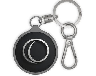 May include: A silver-colored keychain with a round, black pendant featuring a white, crescent-shaped design. The keychain includes a split ring and a lobster clasp for easy attachment to bags or keys.