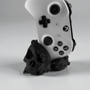 May include: A white Xbox controller is displayed on a black skull-shaped stand. The controller has black buttons and a directional pad. The stand has a detailed skull design with tentacles.