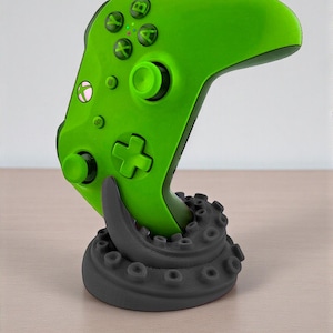 May include: A bright green Xbox controller is displayed on a dark gray octopus tentacle-shaped stand. The controller has black buttons and a directional pad. The stand is on a light brown surface.