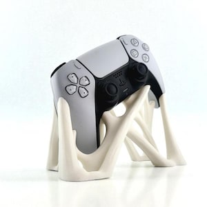 May include: A white and black PlayStation 5 controller resting on a cream-colored, abstract-shaped stand. The controller has a symmetrical design with buttons and joysticks. The stand has a unique, artistic form.