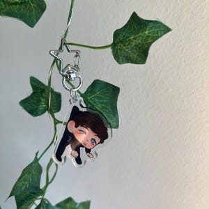May include: A clear acrylic charm featuring a cartoon illustration of a person with brown hair and blue eyes, wearing a black outfit. The charm is attached to a silver star-shaped keychain. Green leaves and vines are in the background.