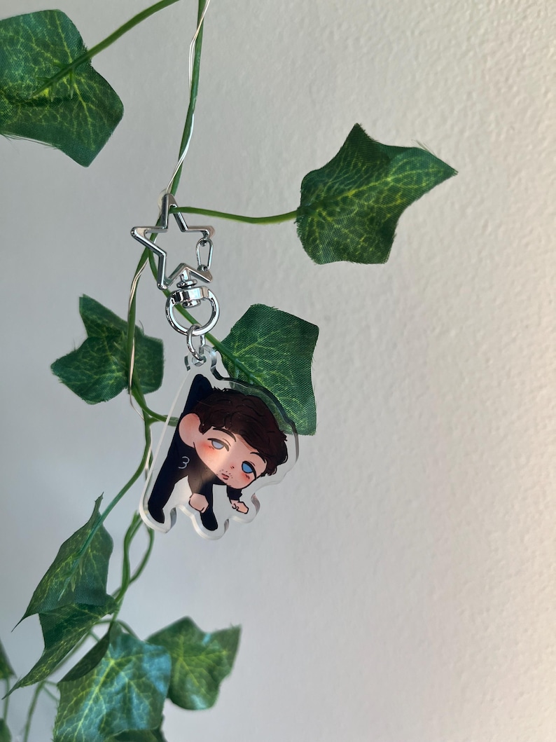 May include: A clear acrylic charm featuring a cartoon illustration of a person with brown hair and blue eyes, wearing a black outfit. The charm is attached to a silver star-shaped keychain. Green leaves and vines are in the background.