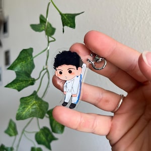 May include: A clear acrylic keychain featuring a cartoon illustration of a person in a white and blue outfit. The keychain has a silver-colored metal ring. The person has black hair and is wearing black shoes. The number 23 is visible on the outfit.