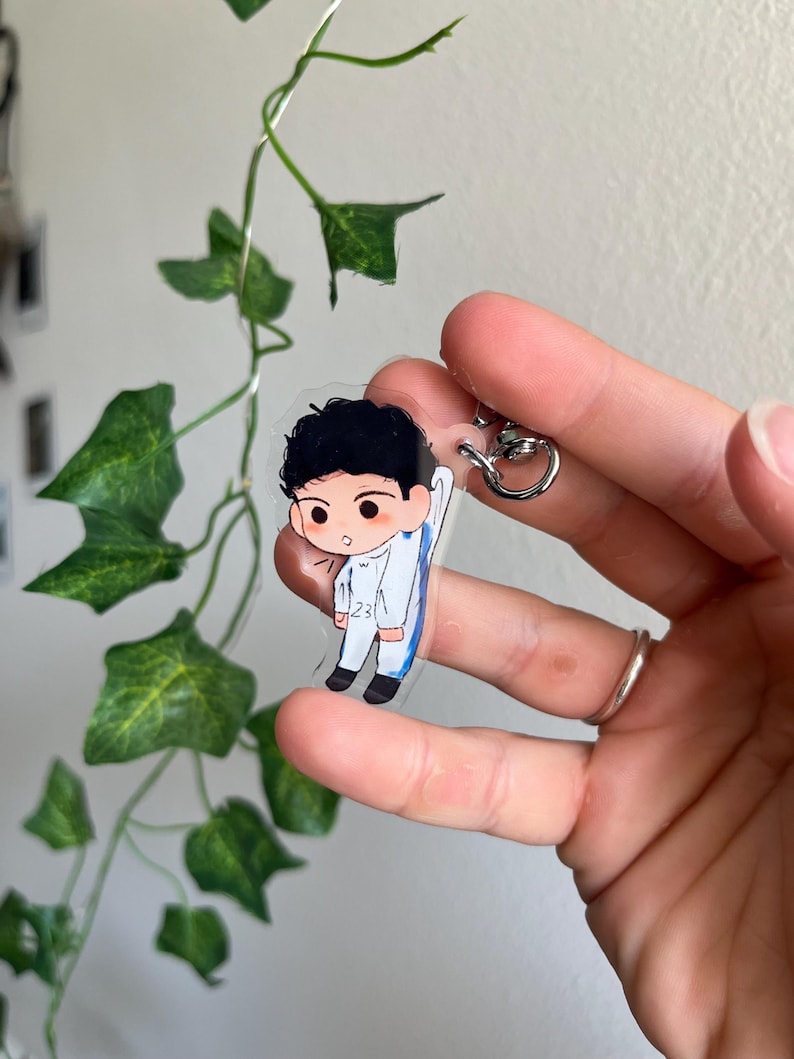 May include: A clear acrylic keychain featuring a cartoon illustration of a person in a white and blue outfit. The keychain has a silver-colored metal ring. The person has black hair and is wearing black shoes. The number 23 is visible on the outfit.