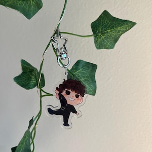 May include: A clear acrylic keychain featuring a cartoon character with brown curly hair, wearing a black outfit with the number 12. The keychain is attached to a silver star-shaped clasp, hanging from a green vine.