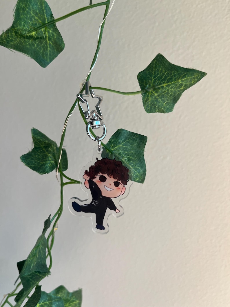 May include: A clear acrylic keychain featuring a cartoon character with brown curly hair, wearing a black outfit with the number 12. The keychain is attached to a silver star-shaped clasp, hanging from a green vine.
