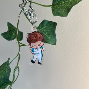 May include: A clear acrylic keychain featuring a cartoon illustration of a person with brown hair in a blue and white outfit. The keychain is attached to a silver star-shaped clasp. Green leaves and vines are in the background.