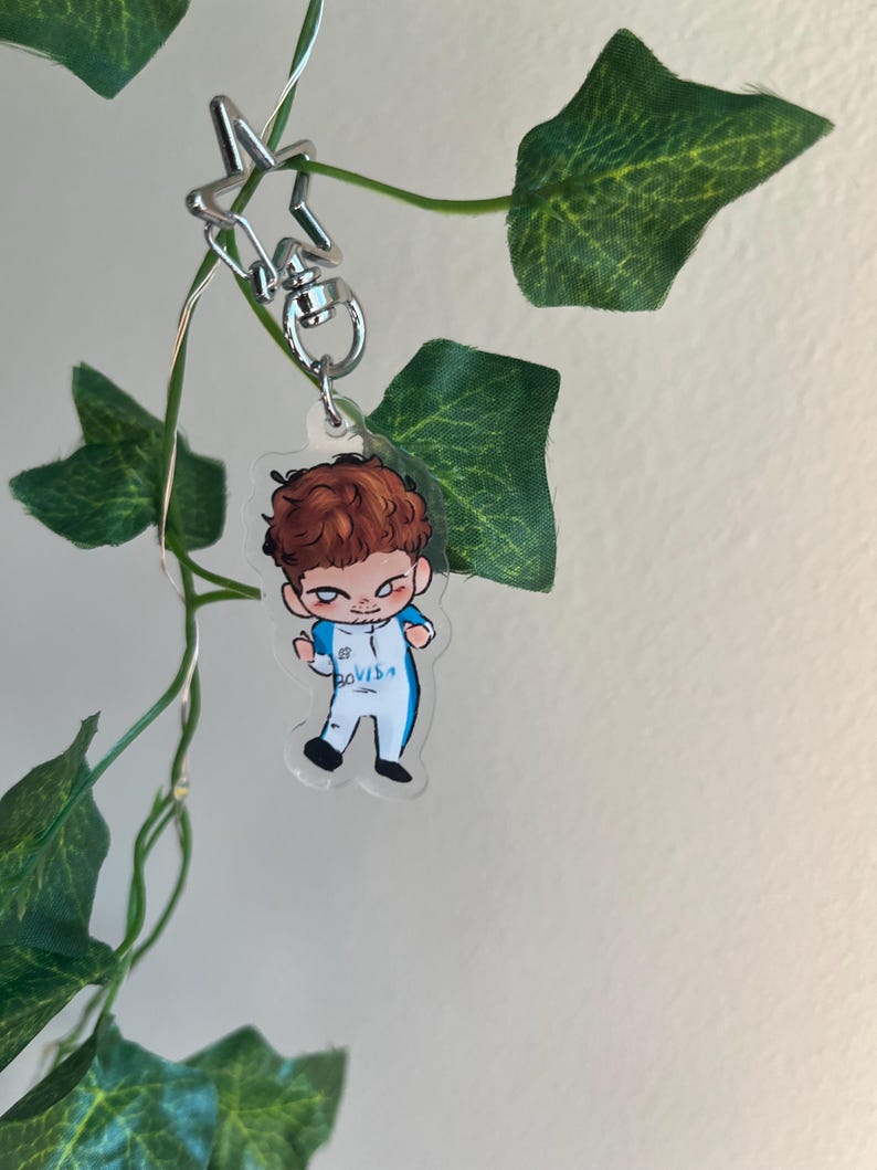 May include: A clear acrylic keychain featuring a cartoon illustration of a person with brown hair in a blue and white outfit. The keychain is attached to a silver star-shaped clasp. Green leaves and vines are in the background.