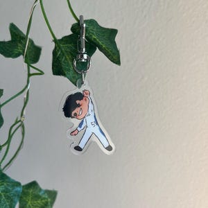 May include: A clear acrylic keychain featuring a cartoon illustration of a person in a white and blue racing suit with the number 55. The keychain is attached to a silver metal clasp and is hanging in front of green leaves.