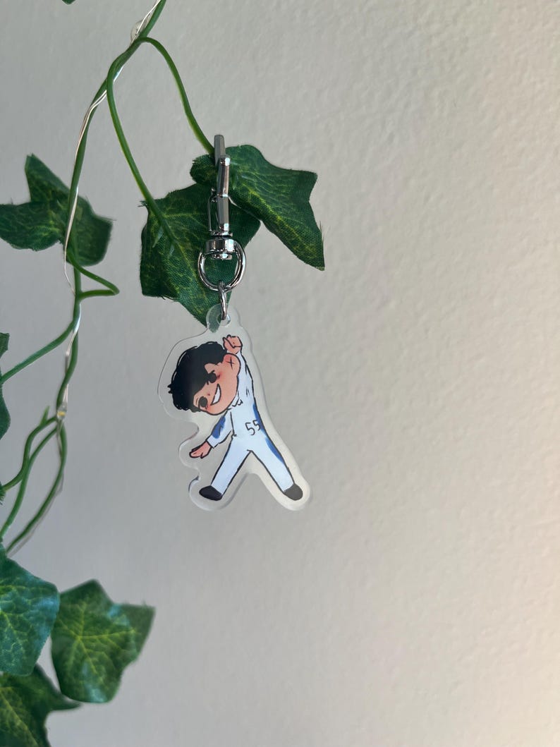 May include: A clear acrylic keychain featuring a cartoon illustration of a person in a white and blue racing suit with the number 55. The keychain is attached to a silver metal clasp and is hanging in front of green leaves.