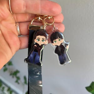 May include: Two clear acrylic keychains featuring cartoon illustrations of characters with black hair, green eyes, and black outfits. The keychains are attached to a gold-colored clasp and a black and gray patterned lanyard.