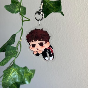 May include: A clear acrylic keychain featuring a cartoon illustration of a person with brown hair and a black and red tracksuit. The keychain is attached to a silver-colored clasp and ring. Green leaves and vines are in the background.