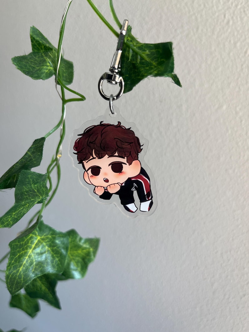May include: A clear acrylic keychain featuring a cartoon illustration of a person with brown hair and a black and red tracksuit. The keychain is attached to a silver-colored clasp and ring. Green leaves and vines are in the background.