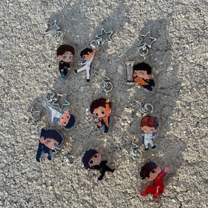 May include: Collection of cartoon character keychains. Each keychain features a small, colorful figure in various poses, attached to a silver star-shaped charm and a keyring. The figures are dressed in different outfits, with varying hair colors and styles.