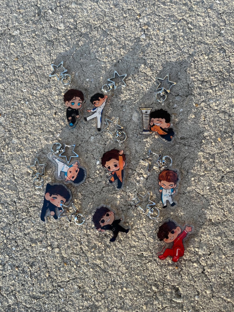 May include: Collection of cartoon character keychains. Each keychain features a small, colorful figure in various poses, attached to a silver star-shaped charm and a keyring. The figures are dressed in different outfits, with varying hair colors and styles.