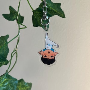 May include: A clear acrylic keychain featuring a cartoon illustration of a person in a white and blue outfit. The keychain is attached to a silver clasp and hangs from a green vine with leaves.