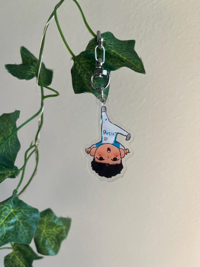 May include: A clear acrylic keychain featuring a cartoon illustration of a person in a white and blue outfit. The keychain is attached to a silver clasp and hangs from a green vine with leaves.