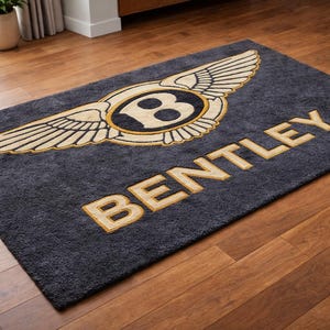 May include: A rectangular, dark gray rug with the Bentley logo in the center. The logo features a gold-colored emblem with wings and the word "BENTLEY" in gold lettering. The rug is placed on a wooden floor.
