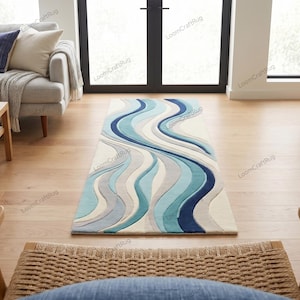 May include: A rectangular area rug with a wavy design in shades of blue, gray, and white. The rug is placed on a hardwood floor in a living room setting, with a sofa, chairs, and a coffee table visible in the background.