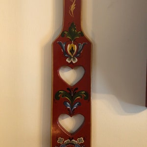 May include: A red wooden paddle with three heart-shaped cutouts and traditional Scandinavian folk art designs painted in blue, green, yellow, and white.