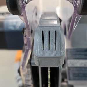 May include: Close-up of a gray power tool component with three vertical slots and a textured surface. The tool has a clear plastic guard and black wheels. The image is a detailed view of a power tool.