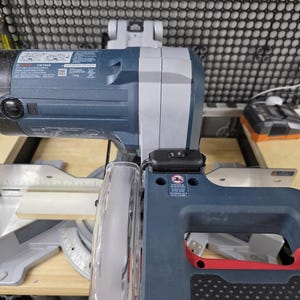 May include: A blue and gray miter saw with a clear blade guard. The saw has a red handle and a black base. The blade is visible, and the saw is set on a wooden surface. The saw is designed for cutting wood.