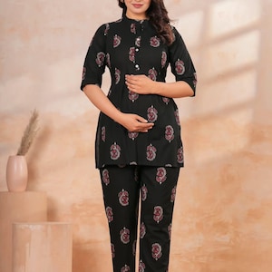 Könnte beinhalten: Ein schwarzes Tunika- und Hosenset mit einem sich wiederholenden Blumenmuster in Rosa, Weiß und Grün. Die Tunika hat einen Stehkragen, eine teilweise Knopfleiste auf der Vorderseite und Dreiviertelärmel, kombiniert mit einer geraden Hose.