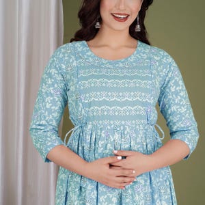 K&ouml;nnte beinhalten: Hellblaues Kleid mit floralem Muster und Dreiviertel&auml;rmeln. Das Kleid hat eine betonte Taille und dekorative wei&szlig;e Spitzendetails am Oberteil. Der Stoff hat ein zartes, flie&szlig;endes Aussehen, das sich f&uuml;r Freizeitkleidung eignet.