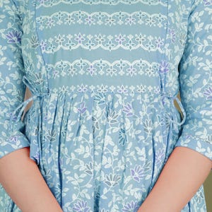 K&ouml;nnte beinhalten: Hellblaues Kleid mit floralem Muster und Dreiviertel&auml;rmeln. Das Kleid hat wei&szlig;e und violette Blumenakzente, einen runden Ausschnitt und eine geschn&uuml;rte Taille mit Bindeb&auml;ndern. Das Gesamtdesign ist feminin und elegant.