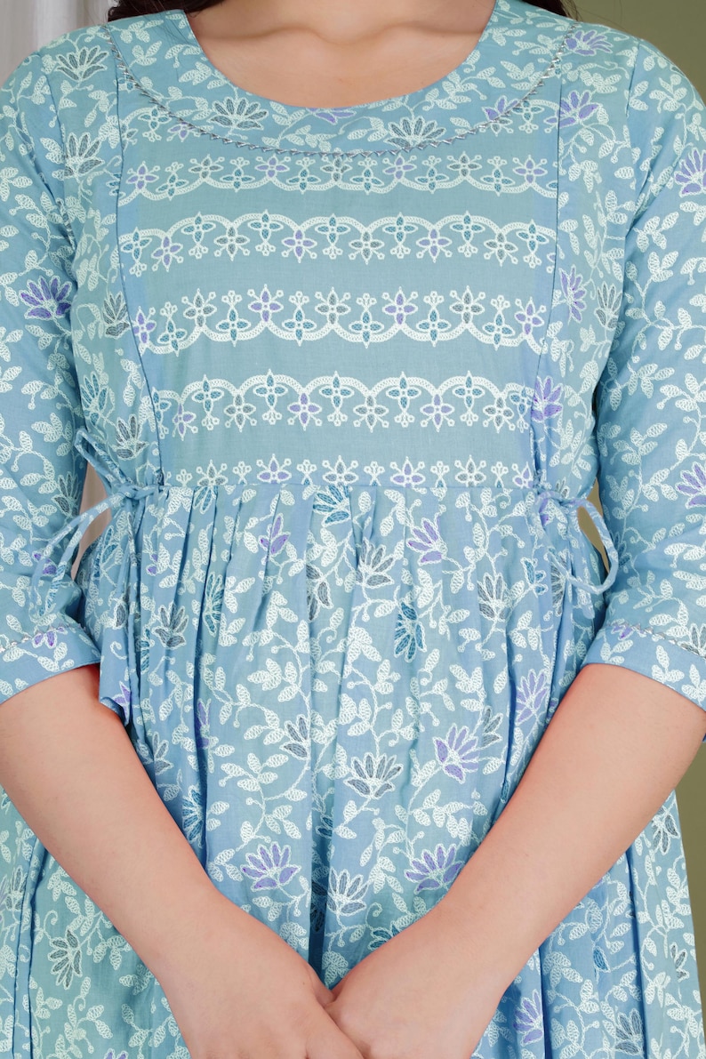 K&ouml;nnte beinhalten: Hellblaues Kleid mit floralem Muster und Dreiviertel&auml;rmeln. Das Kleid hat wei&szlig;e und violette Blumenakzente, einen runden Ausschnitt und eine geschn&uuml;rte Taille mit Bindeb&auml;ndern. Das Gesamtdesign ist feminin und elegant.