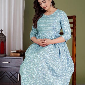 K&ouml;nnte beinhalten: Hellblaues Kleid mit floralem Muster. Das Kleid hat Dreiviertel&auml;rmel und einen runden Ausschnitt. Das Kleid ist in der Taille gerafft und f&auml;llt bis zum Boden. Das Kleid wird von einer Person getragen, die auf einem Holzstuhl sitzt.