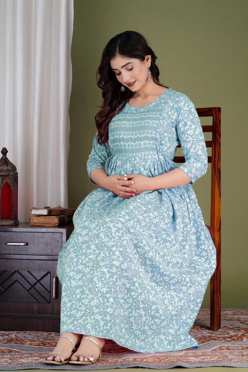 K&ouml;nnte beinhalten: Hellblaues Kleid mit floralem Muster. Das Kleid hat Dreiviertel&auml;rmel und einen runden Ausschnitt. Das Kleid ist in der Taille gerafft und f&auml;llt bis zum Boden. Das Kleid wird von einer Person getragen, die auf einem Holzstuhl sitzt.