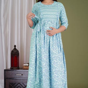 K&ouml;nnte beinhalten: Hellblaues Umstandskleid mit floralem Muster. Das Kleid hat einen runden Ausschnitt, Dreiviertel&auml;rmel und eine geraffte Taille. Das Kleid ist bodenlang und hat einen flie&szlig;enden Rock. Das Model tr&auml;gt goldene Sandalen.