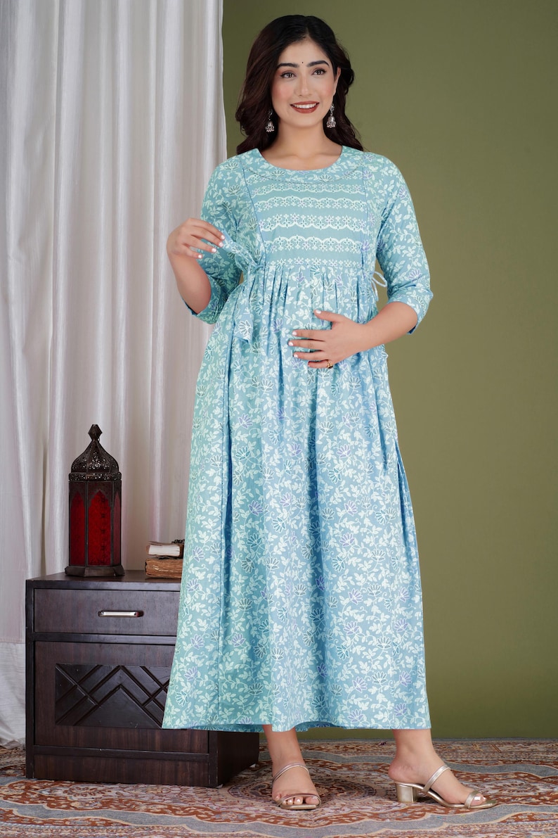 K&ouml;nnte beinhalten: Hellblaues Umstandskleid mit floralem Muster. Das Kleid hat einen runden Ausschnitt, Dreiviertel&auml;rmel und eine geraffte Taille. Das Kleid ist bodenlang und hat einen flie&szlig;enden Rock. Das Model tr&auml;gt goldene Sandalen.