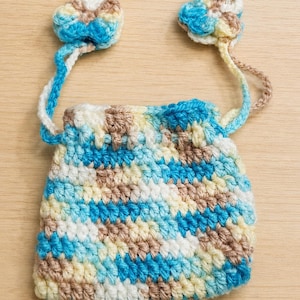 May include: A handmade crochet bag in shades of blue, white, and tan. The bag features a drawstring closure and two flower-shaped accents attached to the straps. The bag is made with a textured stitch.