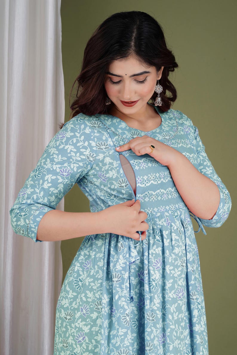 K&ouml;nnte beinhalten: Hellblaues Stillkleid mit floralem Druck und Dreiviertel&auml;rmeln. Das Kleid hat einen runden Ausschnitt und eine geraffte Taille. Das Blumenmuster enth&auml;lt wei&szlig;e und violette Akzente. Das Kleid hat eine diskrete &Ouml;ffnung zum Stillen.