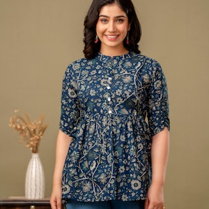 May include: A navy blue top with a floral pattern, featuring a high collar and button-down front. The top has three-quarter sleeves and a gathered waist. The model is wearing blue jeans. A vase with dried flowers is on a wooden table.