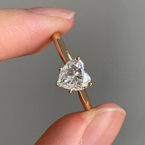 May include: A gold ring featuring a heart-shaped diamond. The diamond is clear and faceted, catching the light. The ring's band is a thin, polished gold. The ring is held up against a neutral background.