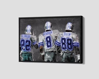 Puede incluir: Obra de arte enmarcada que presenta a tres jugadores de fútbol americano con uniformes blancos y azules, vistos por la espalda. Los jugadores llevan cascos y camisetas con los nombres L. SMITH, AIKMAN e IRVIN, y los números 22, 8 y 88. El fondo es una superficie oscura y texturizada.