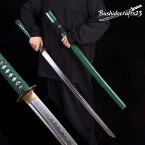 Puede incluir: Una espada katana con una vaina verde texturizada y un mango verde a juego con detalles en forma de diamante blanco. La hoja tiene una línea de hamon ondulada visible. El texto Bushidocrafts23 aparece en la esquina superior derecha.