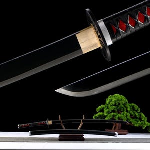 Può includere: Una spada katana nera con una lama curva, con un'impugnatura nera con accenti rossi a forma di diamante. La spada poggia su un supporto di legno, con un fodero nero sottostante. Un piccolo bonsai è sullo sfondo.