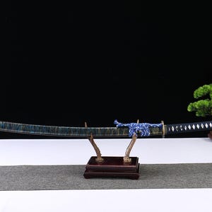 May include: A Japanese katana sword with a blue and black blade, displayed on a wooden stand. The handle is black with white diamond-shaped accents. A blue and white cord wraps the handle. A small bonsai tree is in the background.