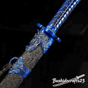 Könnte beinhalten: Nahaufnahme eines dekorativen Schwertes in Blau und Silber. Das Schwert hat detaillierte Drachenmotive auf dem Griff und der Scheide. Die Klinge ist leuchtend blau und die Scheide dunkelgrau mit geprägten Mustern. Der Text "Bushidocrafts23" ist sichtbar.