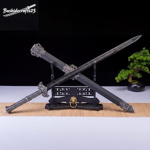 May include: A black sword with an ornate handle and blade, resting on a wooden stand. The sword's blade has a detailed etched design. The stand features a lion's head and ring. The brand name "Bushidocrafts23" is visible in the top left corner.