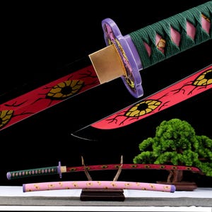May include: A decorative katana sword with a red blade featuring yellow eye designs. The hilt is green and purple, and the scabbard is pink with yellow floral patterns. The sword is displayed on a wooden stand against a black background.