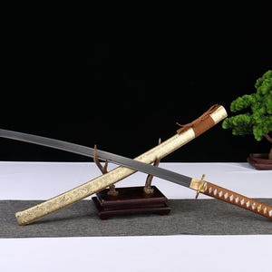 May include: A Japanese katana sword with a black blade and a decorative gold sheath. The handle is wrapped in a brown pattern. The sword rests on a wooden stand, with a bonsai tree in the background.