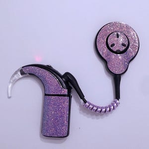May include: A purple glitter hearing aid with a black accent and a clear ear piece. The hearing aid is connected to a round, matching purple glitter component with a black button. The hearing aid has a small red light.