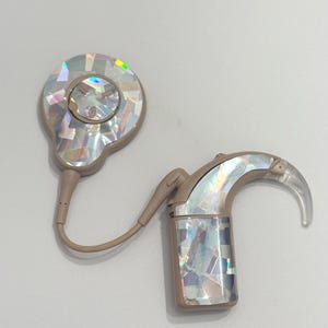 Könnte beinhalten: Ein beigefarbenes Cochlea-Implantat mit schillernden, mosaikartigen Details. Das Gerät besteht aus zwei Hauptkomponenten, die durch einen dünnen, beigefarbenen Draht verbunden sind. Die größere Komponente ist oval, während die kleinere eine gebogene Form mit einer transparenten Spitze hat.