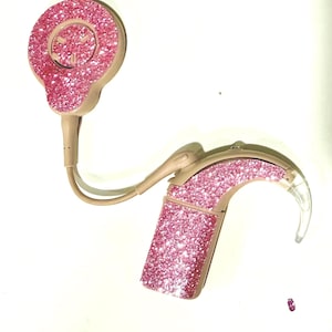 May include: A pair of hearing aids with a pink glitter finish. The hearing aids have a beige base color and a pink glitter coating on the main components. The design includes a circular piece and a curved piece that fits over the ear.