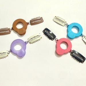 May include: Four hair accessories, each featuring a circular, solid-colored plastic charm in brown, light blue, purple, or pink. Each charm is attached to two metal snap-style hair clips by small silver-toned jump rings for secure attachment.